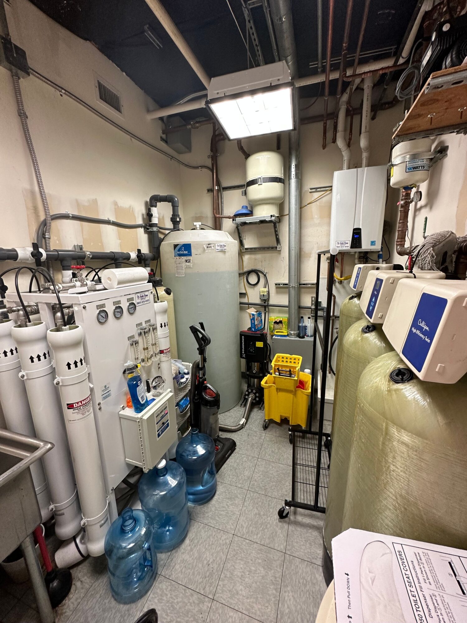 Small utility room with water filters, tanks, and equipment, including water bottles on the floor, pipes, and a ceiling light.