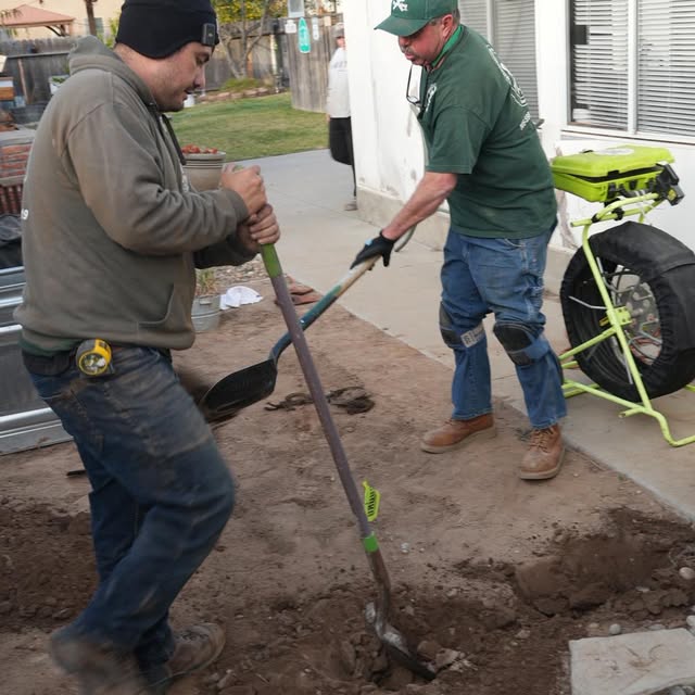 Two men dig in soil with shovels on a sidewalk, one wearing a gray hoodie and the other a green jacket.