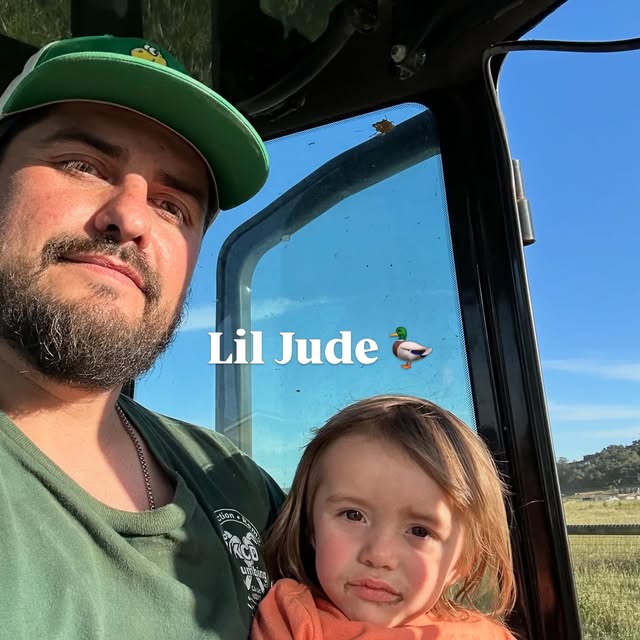 A man and a young girl inside a vehicle with a blue sky and field outside. The man wears a green cap and shirt. The girl looks at the camera with a neutral expression.