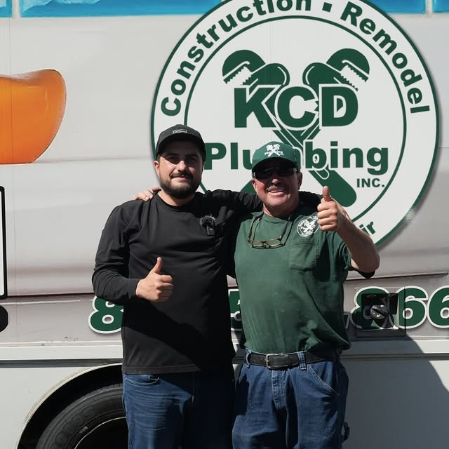 Two men standing in front of a company sign, smiling and giving thumbs-up gestures.
