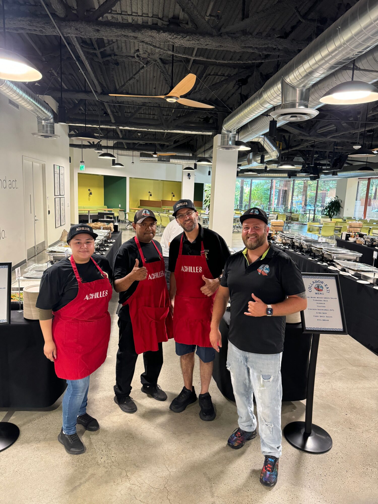 Four people standing inside a restaurant, three wearing red aprons and one in a black shirt, smiling and posing for the photo.