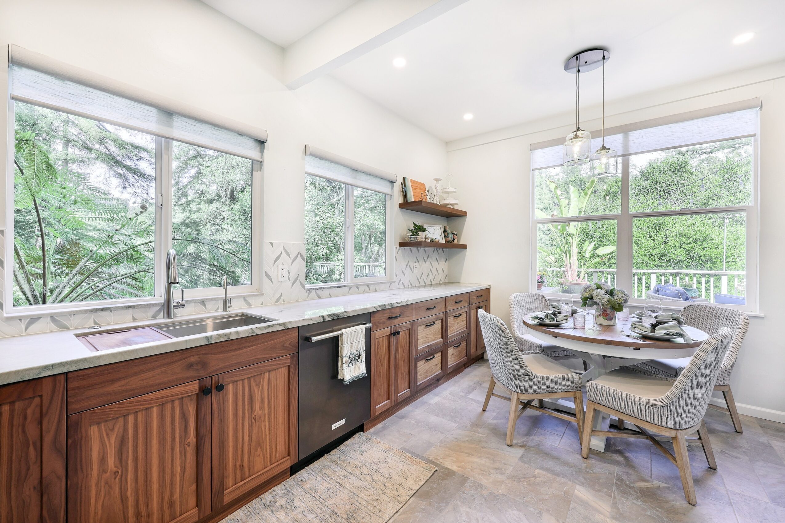 Bright kitchen with large windows, wooden cabinets, round dining table with four chairs, and a view of greenery outside.