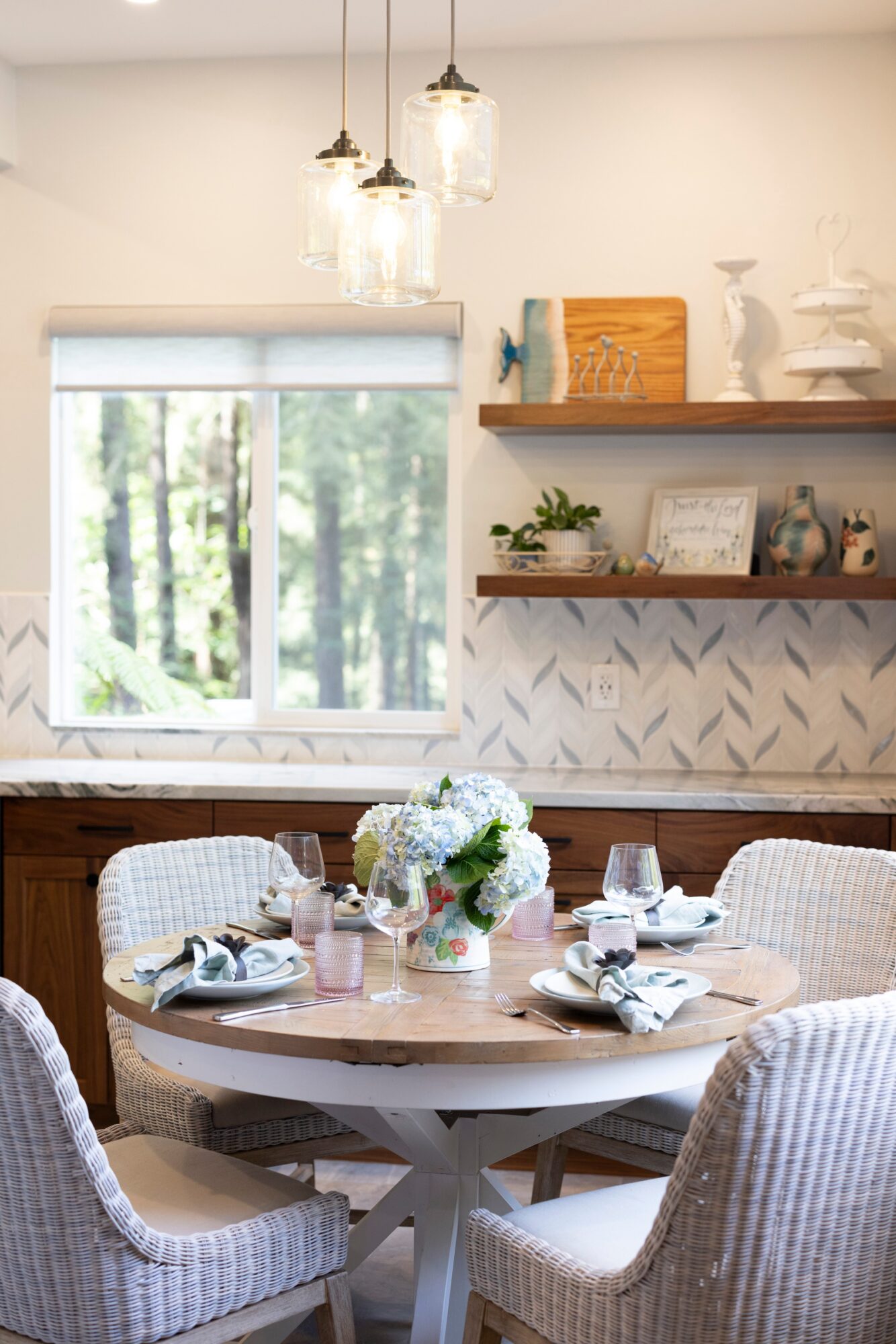 Dining table set with plates, glasses, and a floral centerpiece in a bright room with a window and decorative shelves.