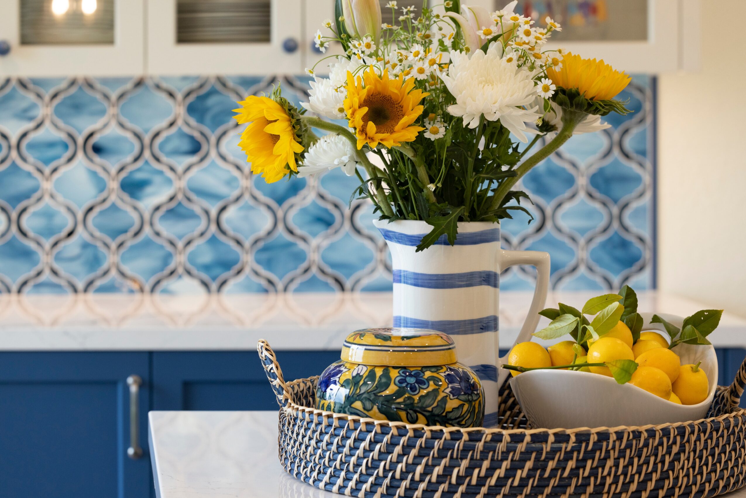 Vase with sunflower and white flowers on kitchen counter, with bowl of lemons nearby, blue patterned backsplash in background.