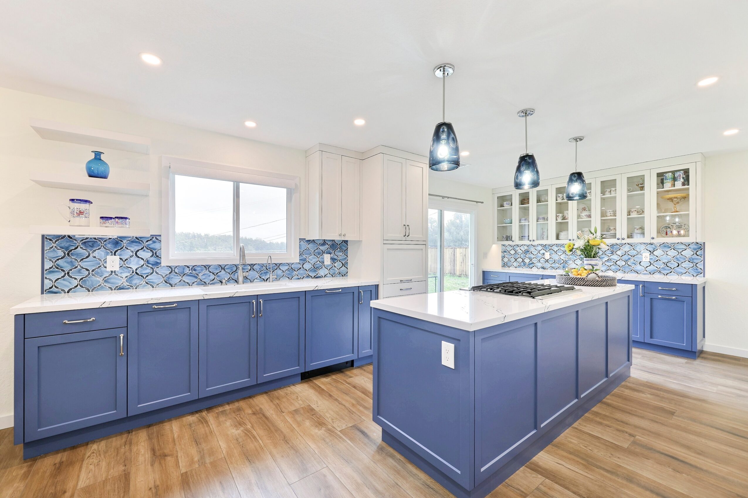 Modern kitchen with blue cabinets, white countertops, and a central island, illuminated by pendant lights, with hardwood floors.