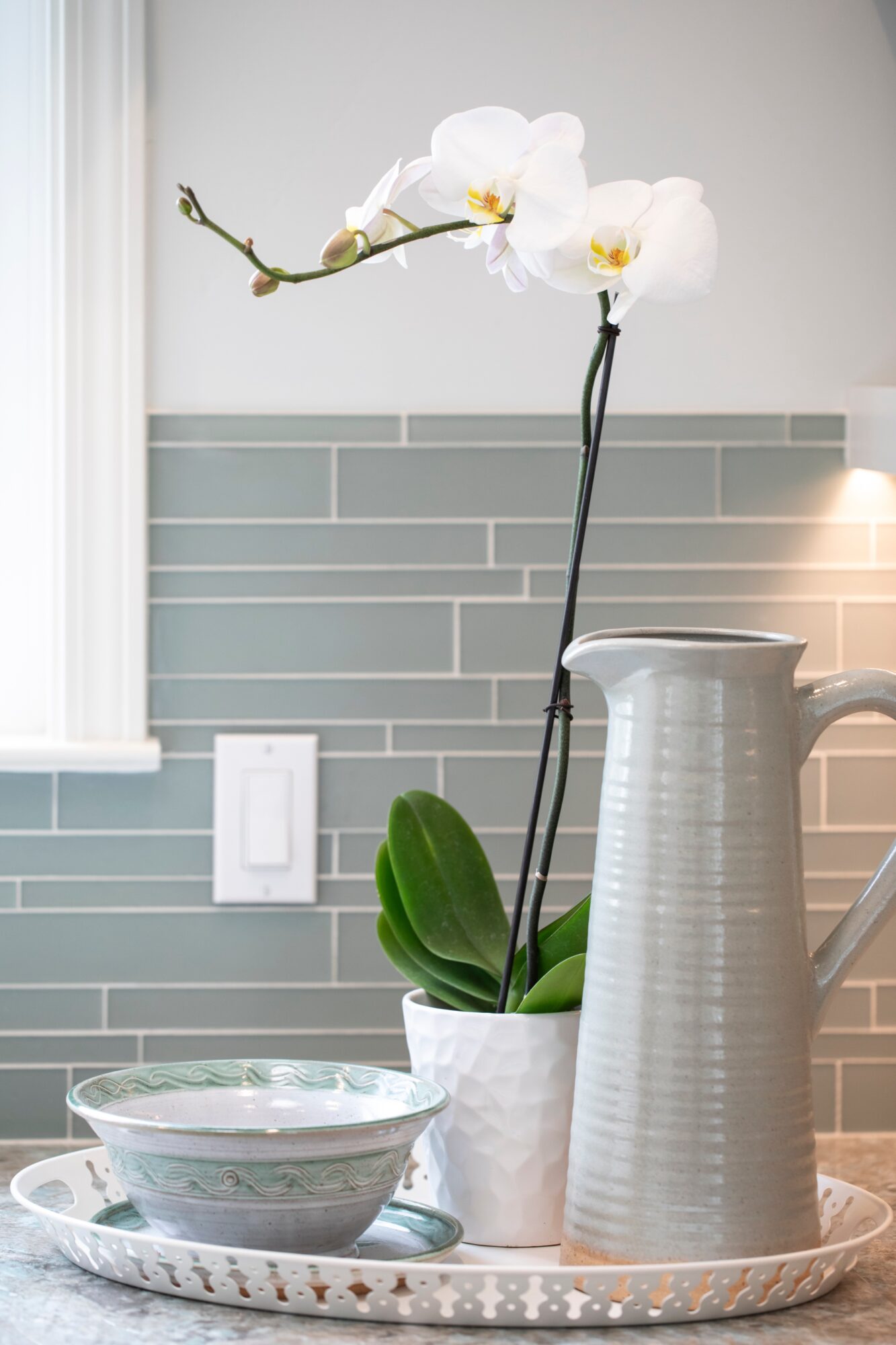White orchid in a pot, tall white pitcher, bowls on a tray, kitchen backsplash, window, and light switch.