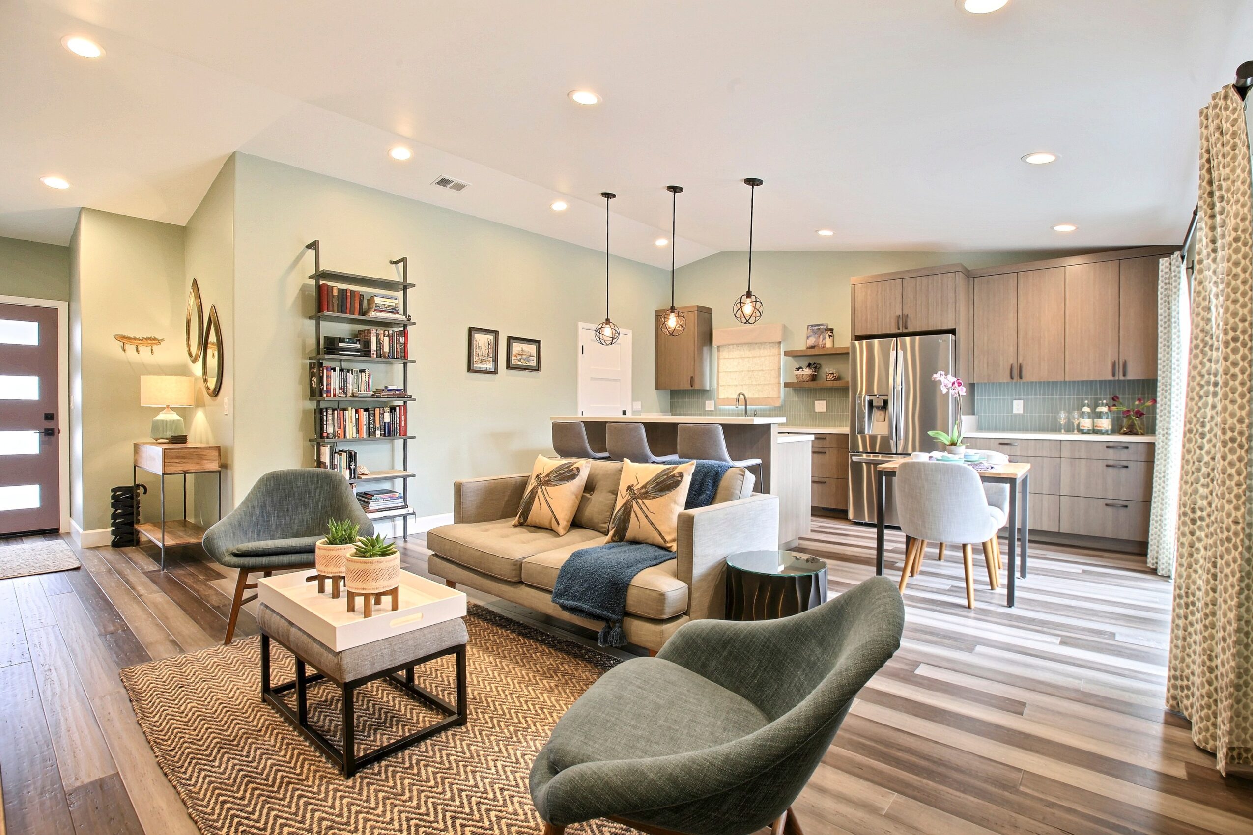 Living room with sofa, armchair, bookshelf, and open kitchen area, with natural light and modern decor.
