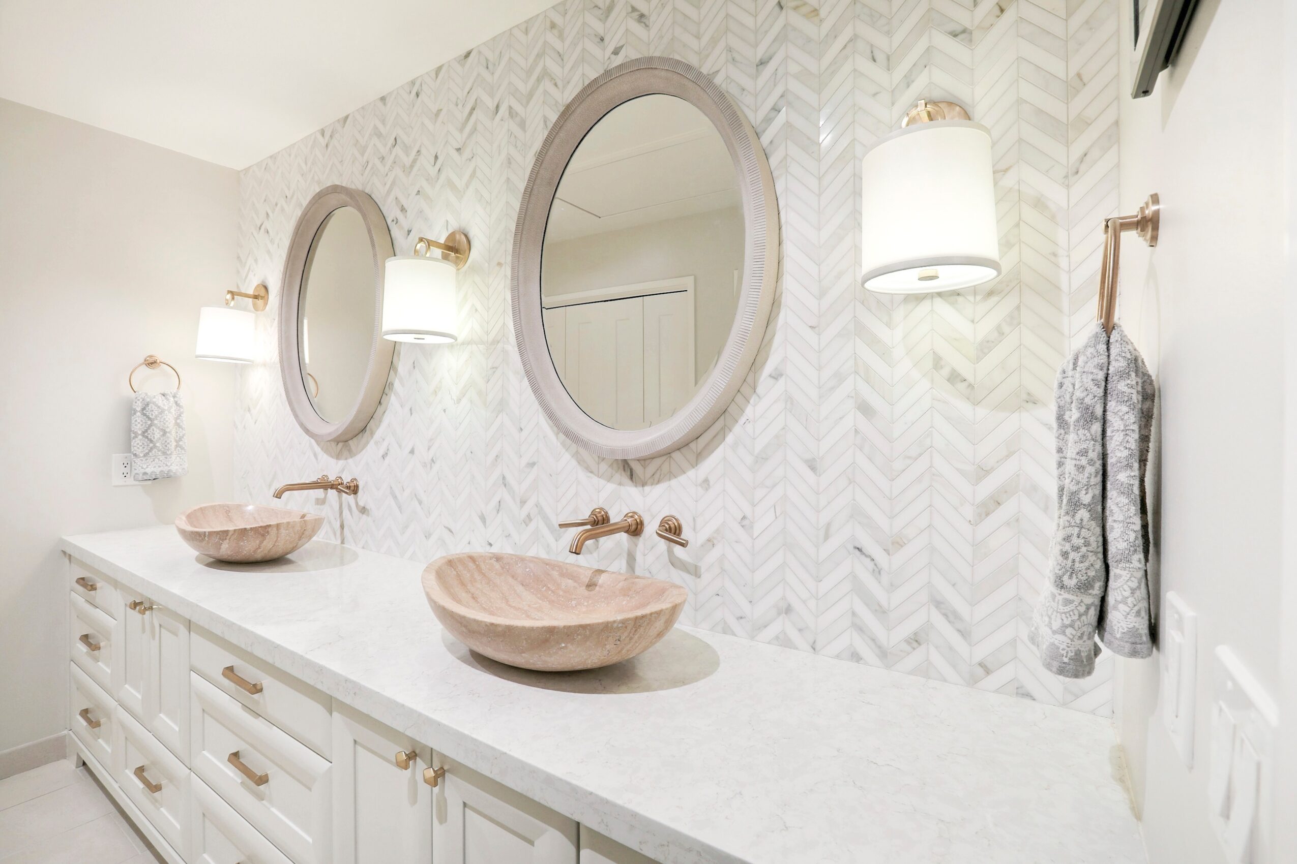 Bathroom with two oval mirrors, two wall-mounted lights, and a white vanity with two vessel sinks, patterned wall tiles, and a towel.