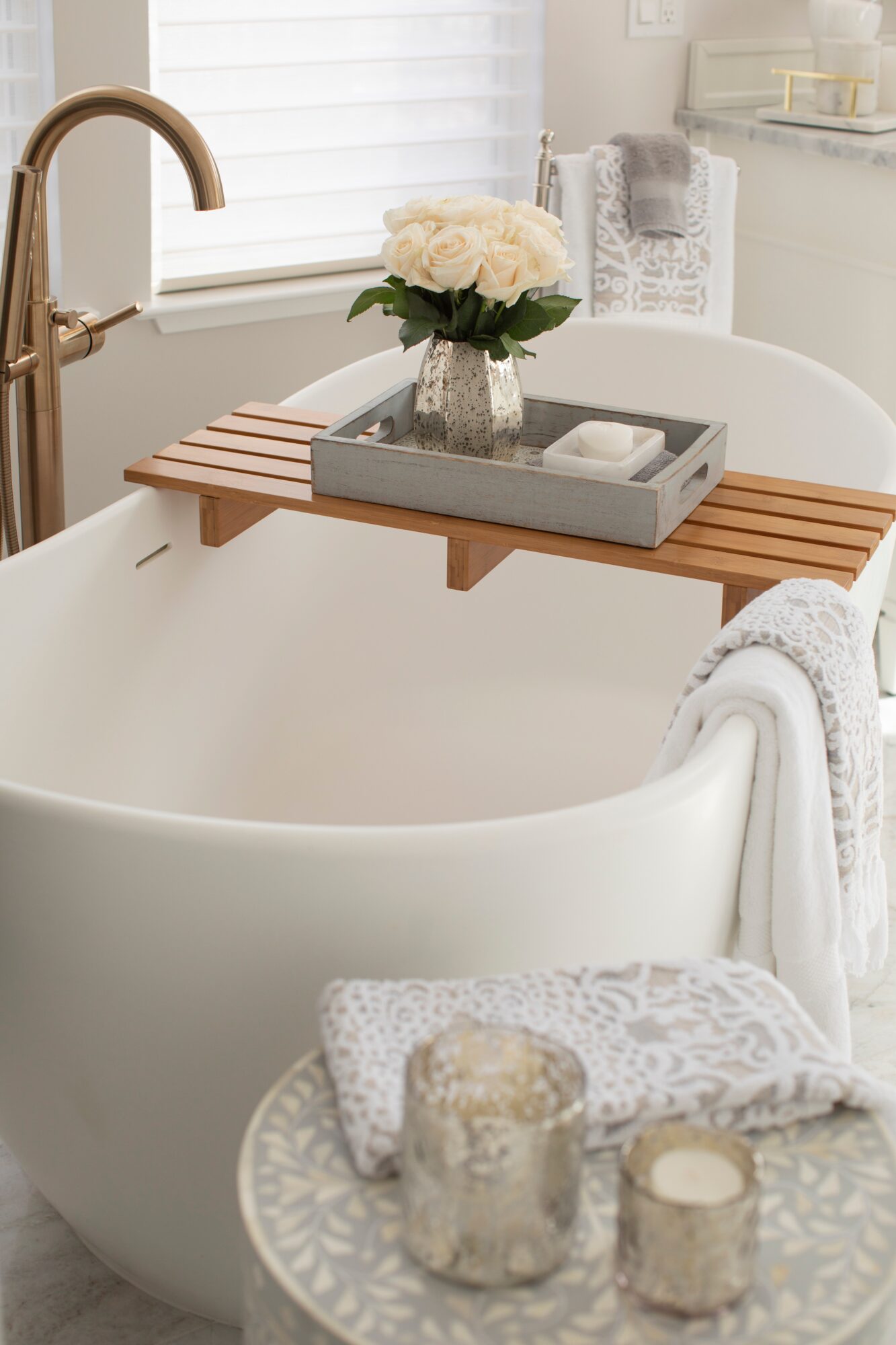 Bathtub with a wooden tray holding a vase of white roses, in a bright bathroom with white decor.