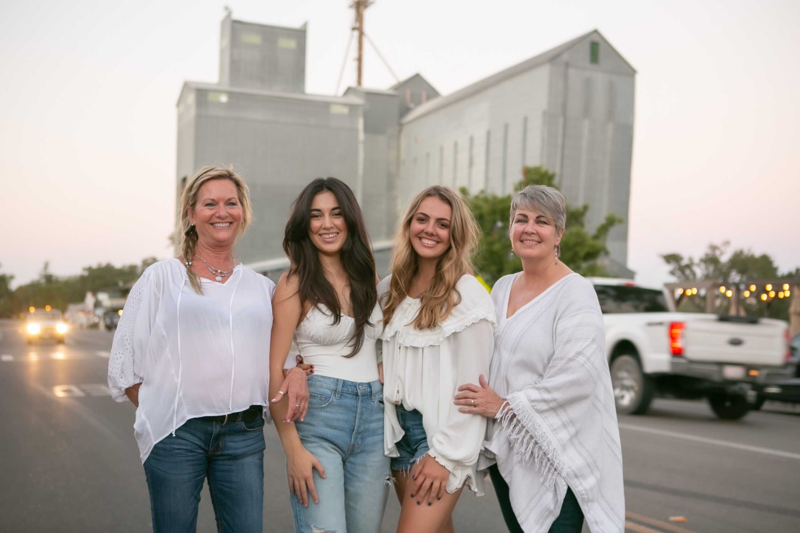Four women standing together outdoors, smiling, with buildings and cars in the background.