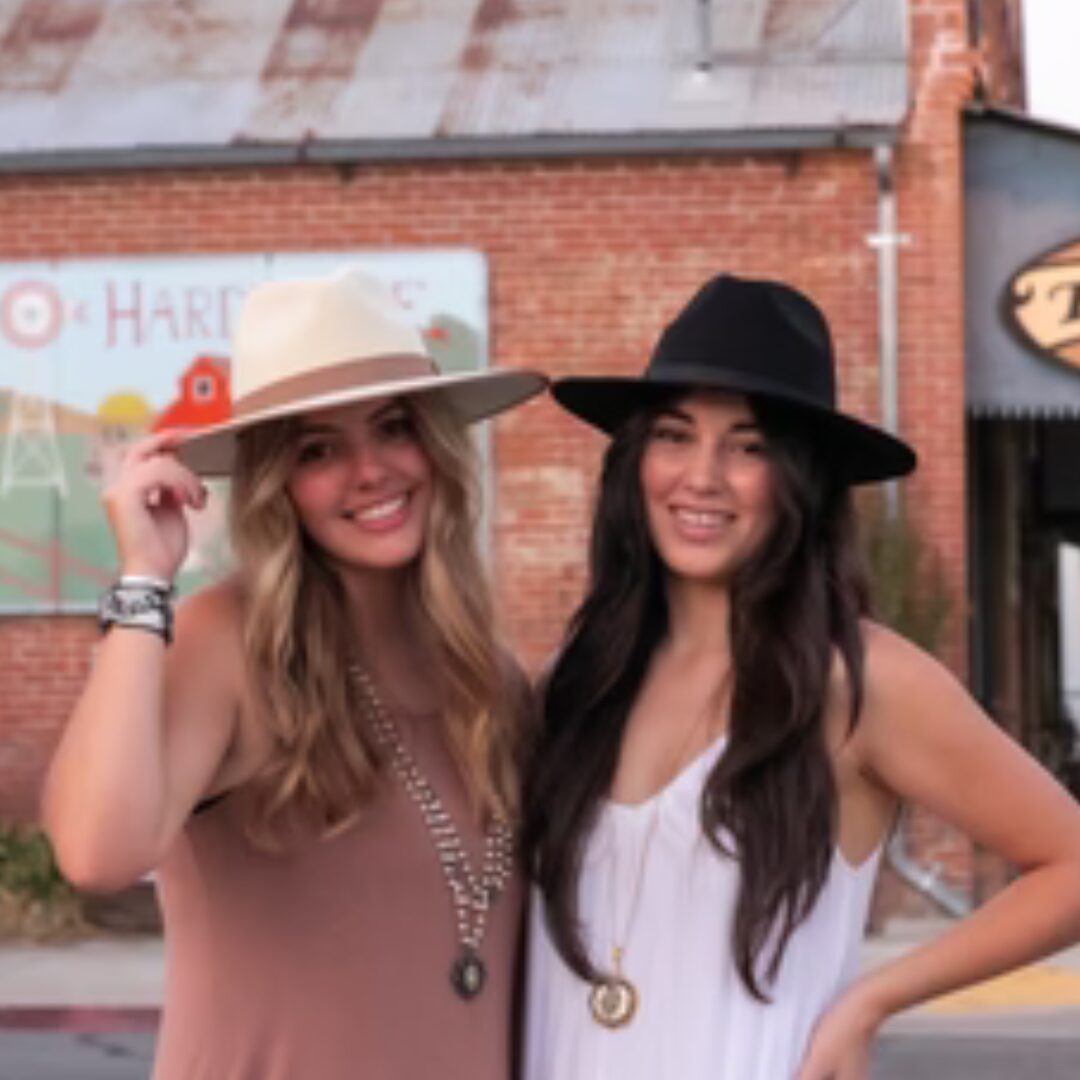 Two women smiling, wearing wide-brimmed hats, standing outdoors in front of a brick building with a colorful sign.