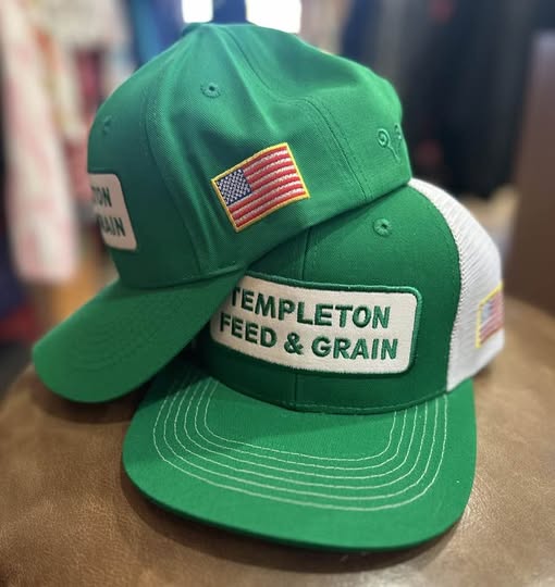 Two green baseball caps with patches, one displaying an American flag and text, the other with text about feed and grain, on a surface.