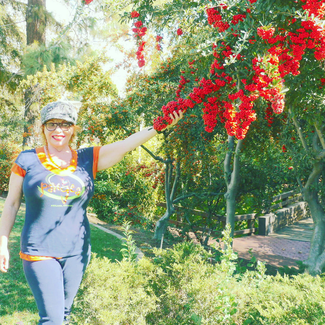Woman in sunglasses and hat reaching for red flowers on a tree in a sunny park.