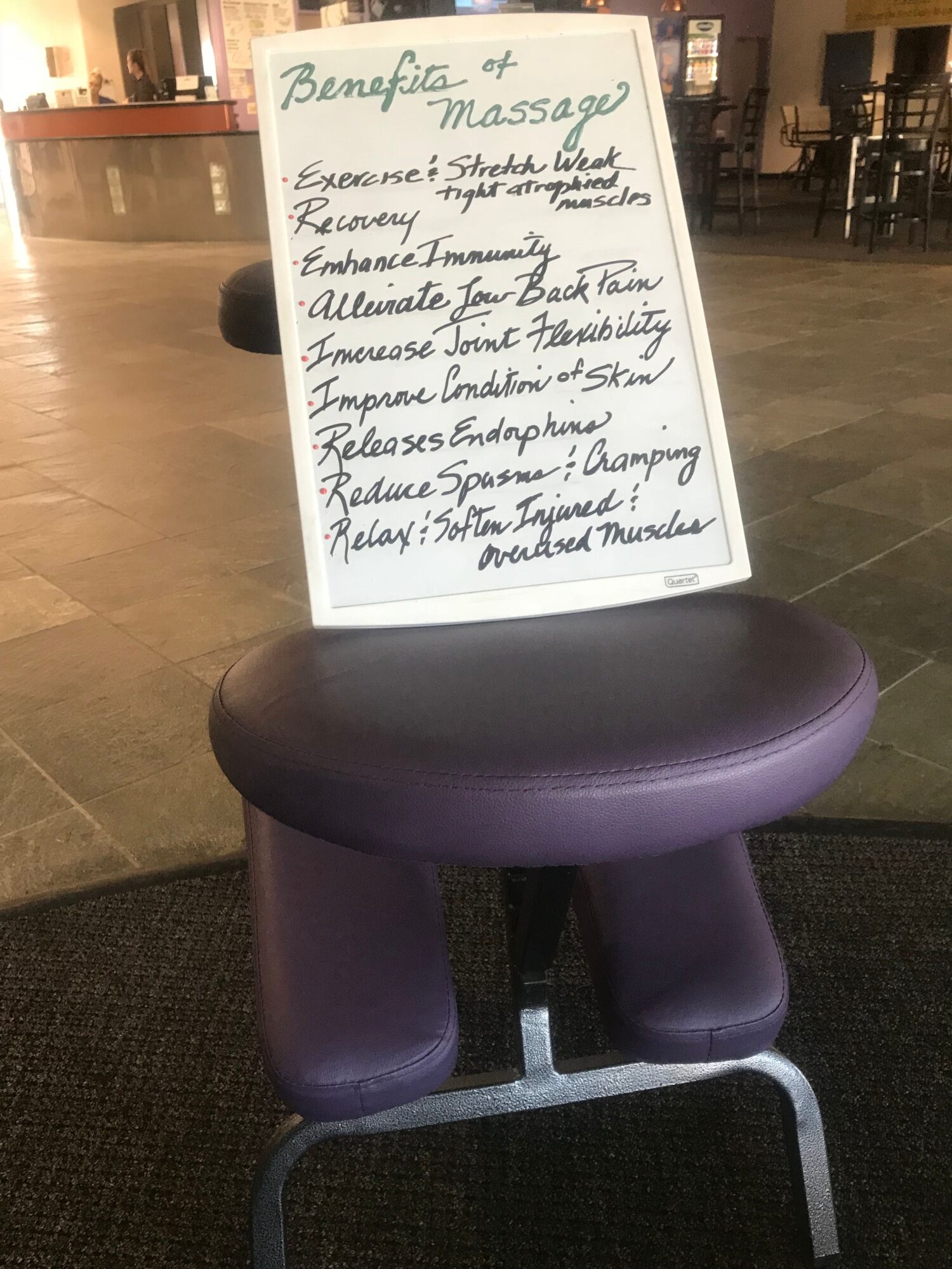 A purple massage chair with a whiteboard sign in front of it, on a wooden floor in a room with shelves.