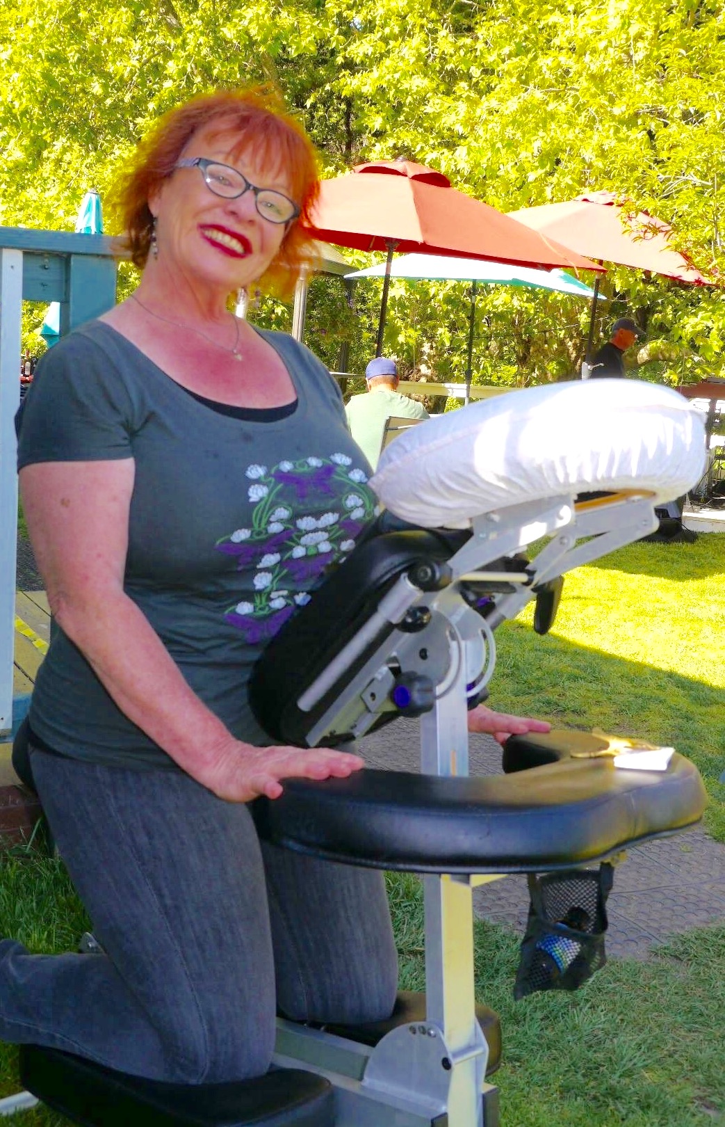 Woman with red hair and glasses smiling outdoors, standing next to a massage table, with trees and a red umbrella in background.