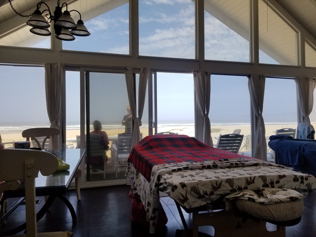 Interior room with large windows showing beach and ocean view, bed with red blanket, and chairs outside on balcony.