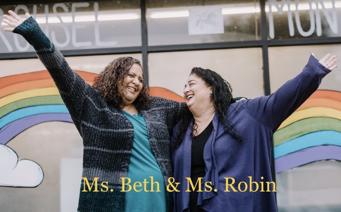 Two women with arms raised, smiling, standing in front of a rainbow mural with rainbow and cloud illustrations.
