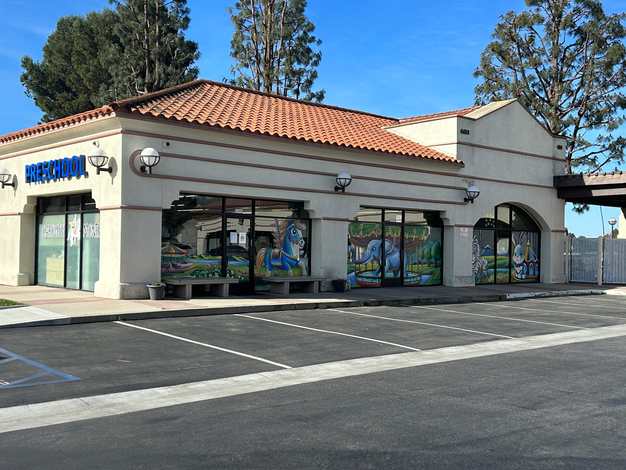 Single-story building with a red-tiled roof, large windows with murals, and a parking lot in front.