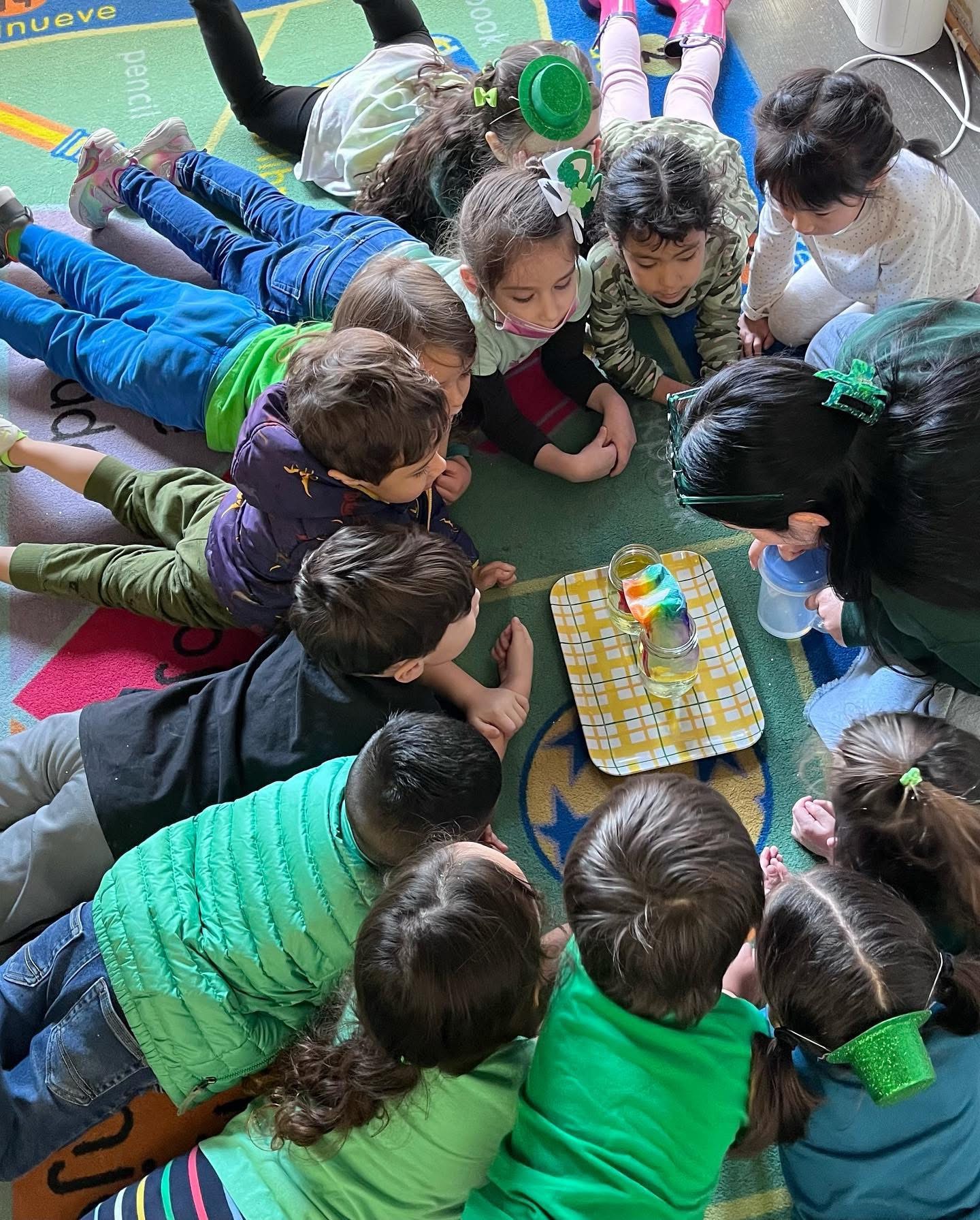 Children gather around a table, engaging with a colorful object, on a green floor with various items nearby.
