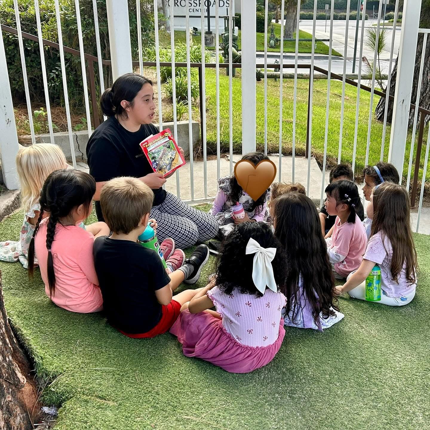 Adult woman reading a book to a group of children sitting on artificial grass outside, behind a white fence.