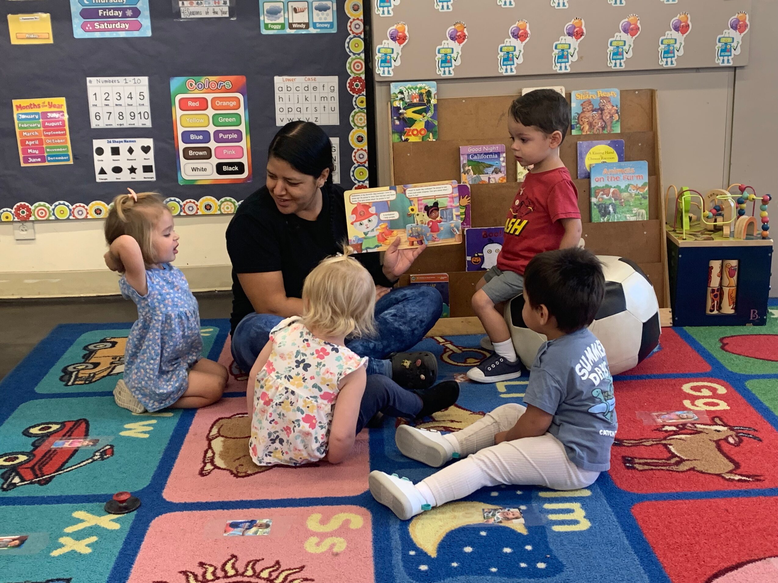 Teacher reading a book to four children sitting on a colorful classroom carpet with educational decorations.