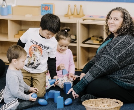 Group of children and a woman playing with blue blocks on the floor in a classroom.