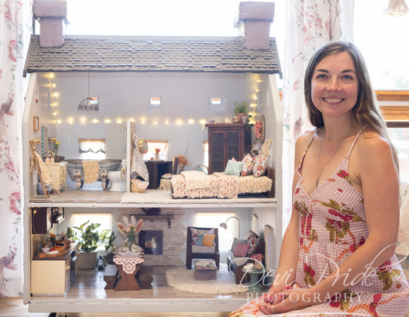Woman smiling in front of a detailed dollhouse with furniture and decorations, indoors with curtains and natural light.