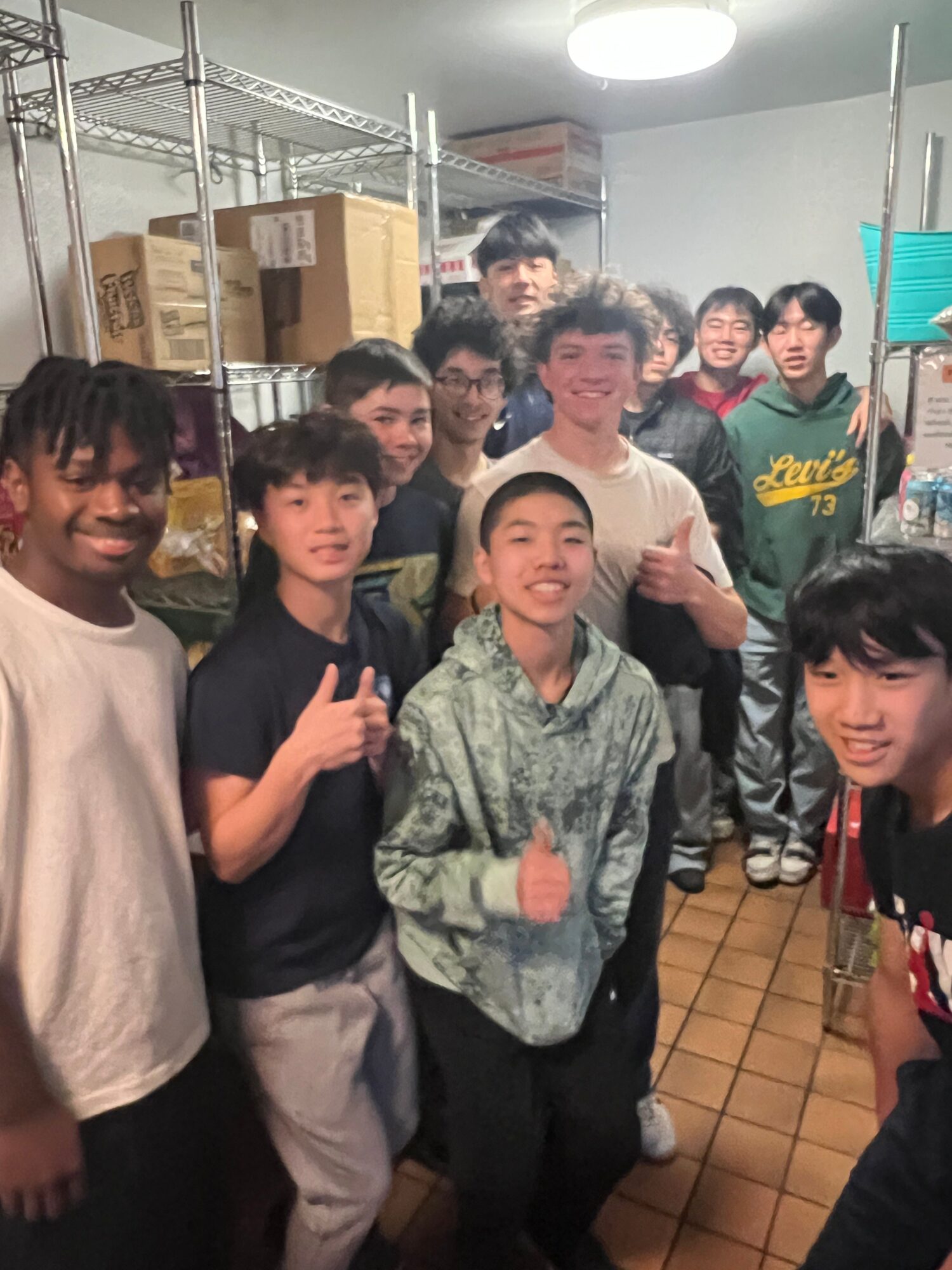 Group of young people in a storage room with shelves and boxes, smiling and giving thumbs up.