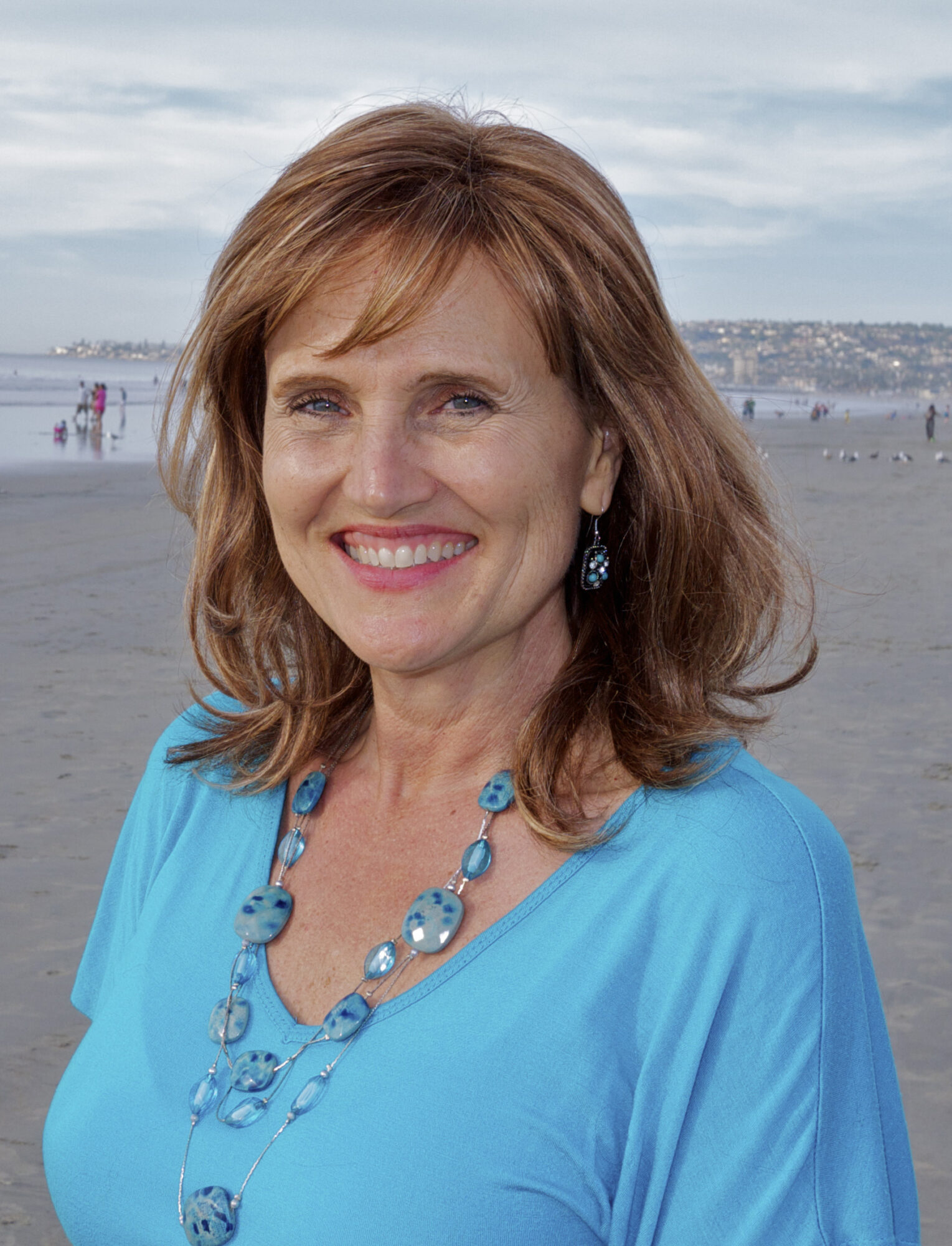 Woman with shoulder-length brown hair smiling, wearing a blue top and jewelry, standing outdoors near water.