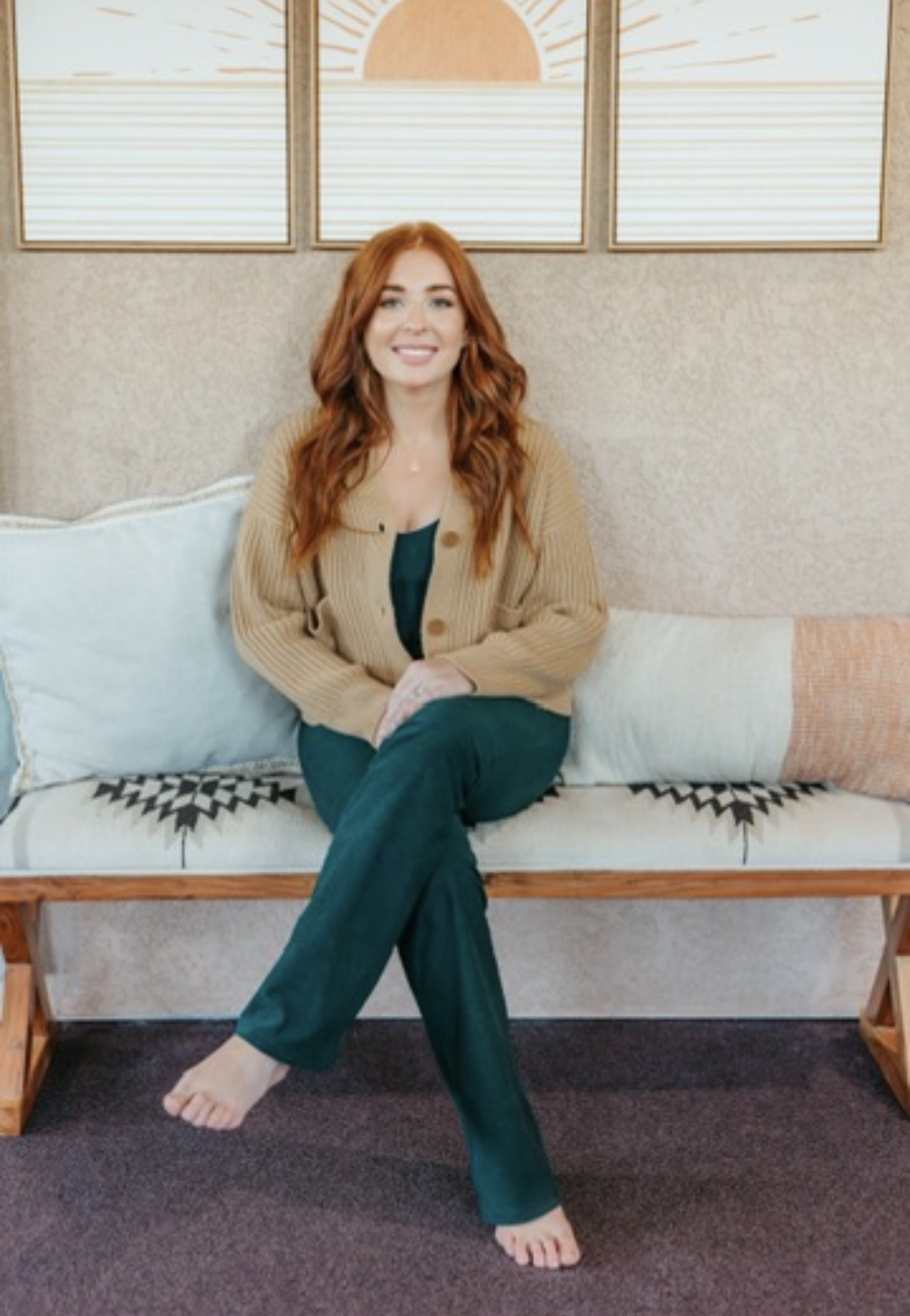 Woman with long red hair sitting on a bench, smiling, wearing a beige jacket and teal pants, in a room with windows.