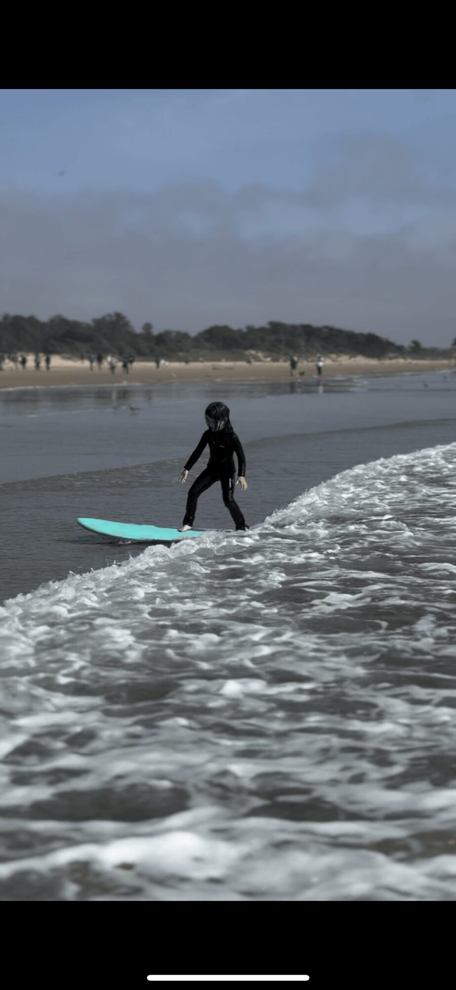 Child in wetsuit standing on surfboard in ocean waves near sandy beach with trees in background.