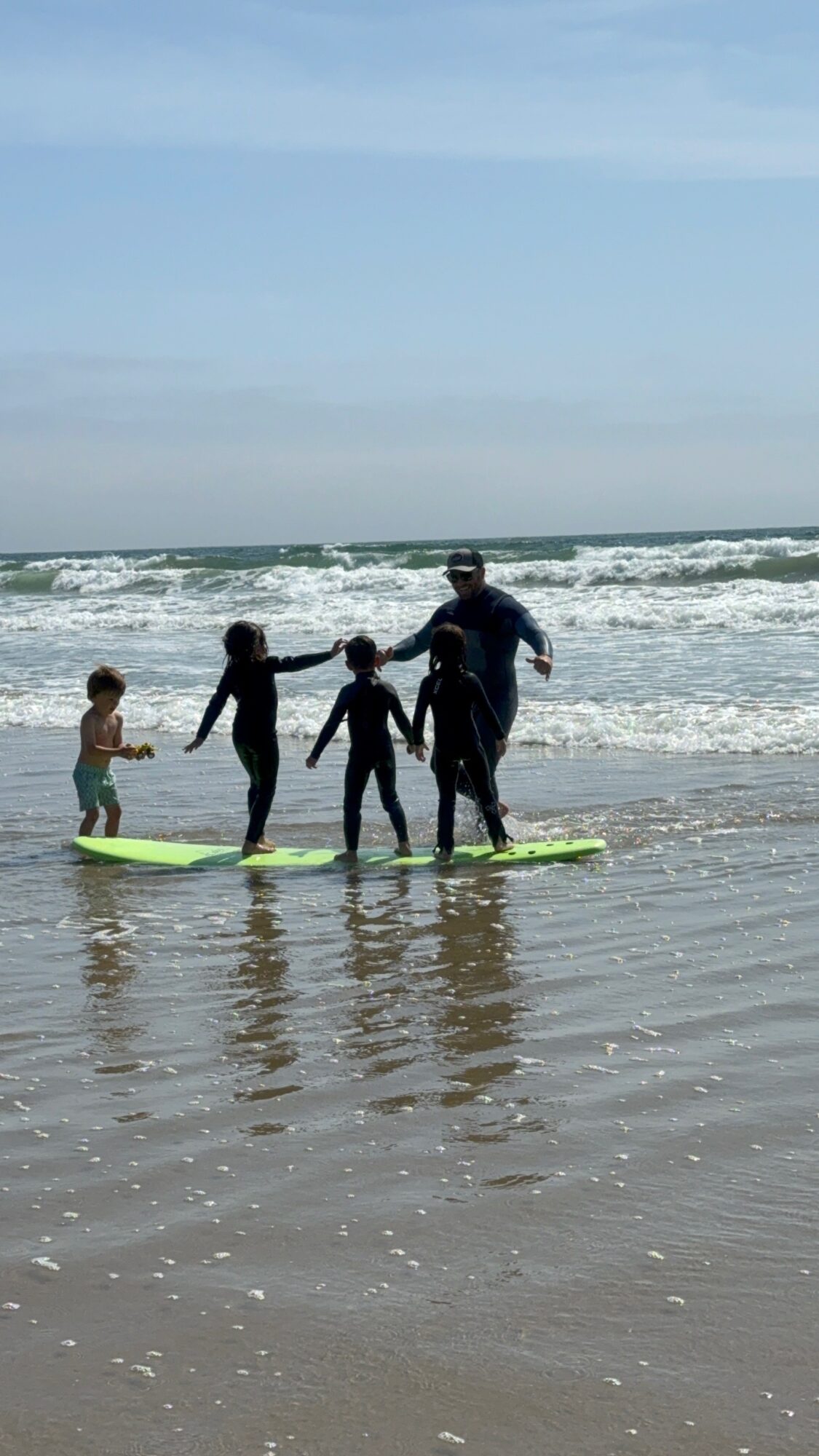 Four people, including children, stand on surfboards in shallow water at the beach, holding hands, with waves and sky in background.