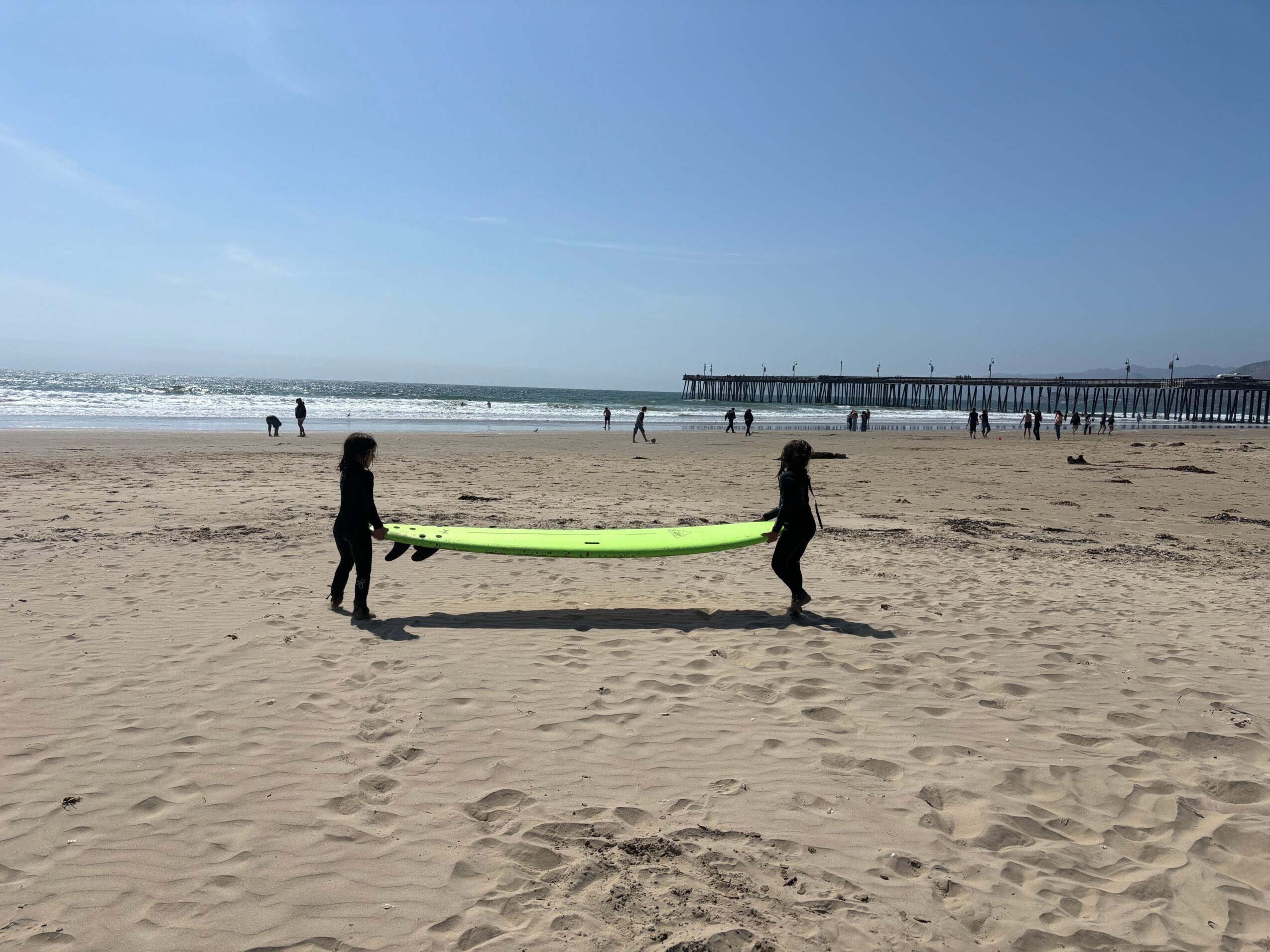 Two children hold a bright green surfboard on a sandy beach with people and a pier in the background.