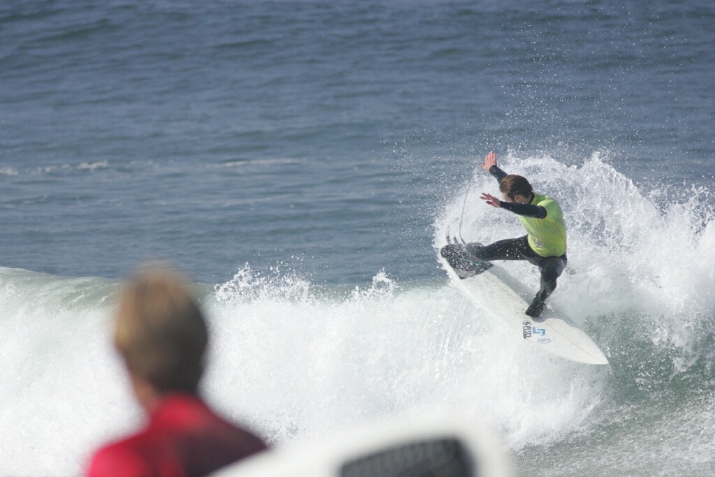 Person surfing on a wave with another person watching in the foreground, ocean background, bright day.