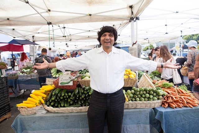 Person with arms outstretched standing in front of a vegetable stall at an outdoor market, smiling.
