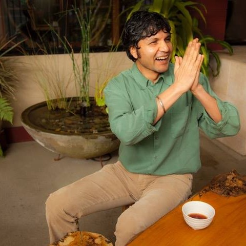 Person with short dark hair, wearing a green shirt, smiling with hands pressed together, sitting at a table with a bowl.