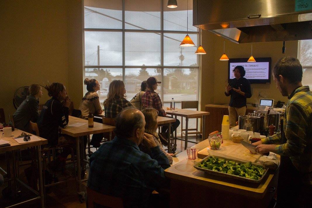 People seated at tables in a cafe or restaurant, listening to a person giving a presentation or speech, with large windows in the background.