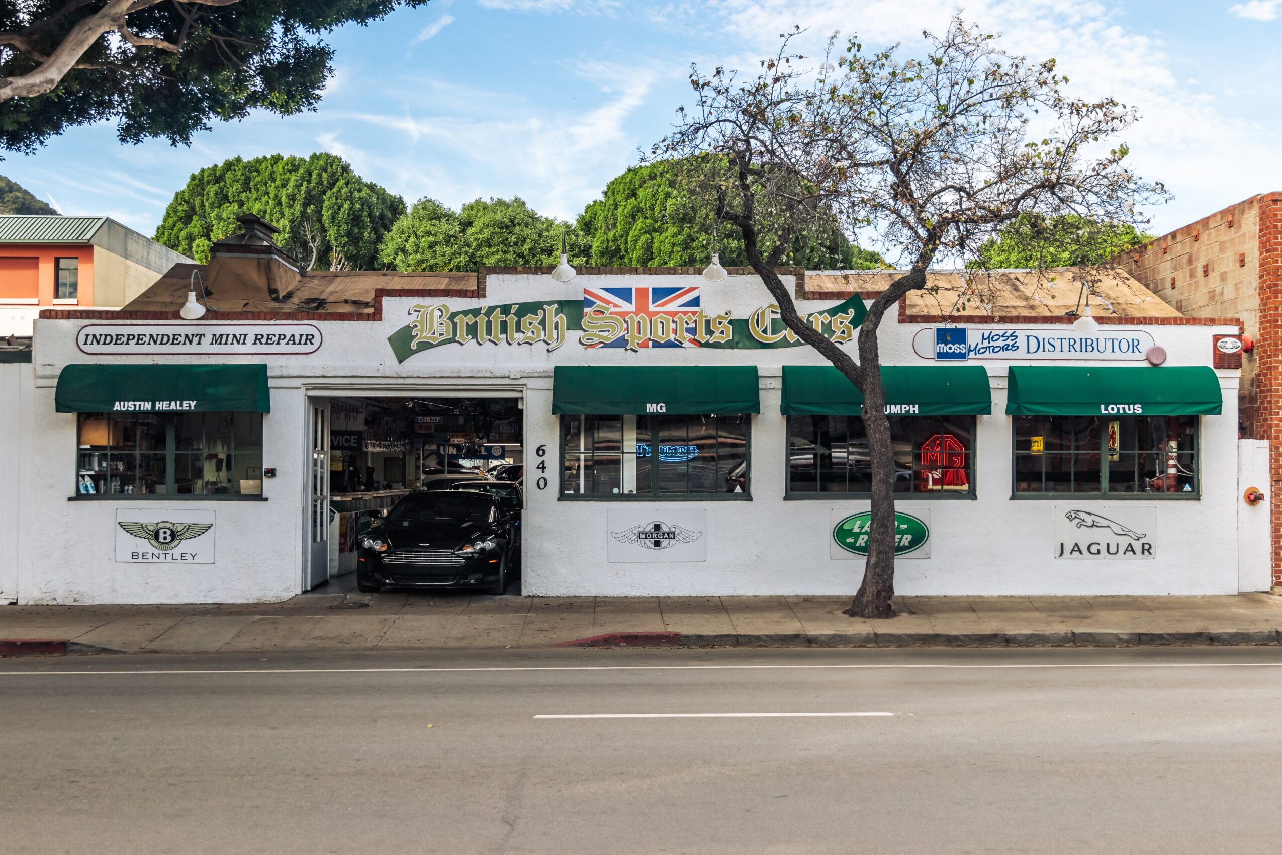 Auto repair shop with cars inside and outside, signage for Jaguar and Land Rover, trees in front, blue sky.