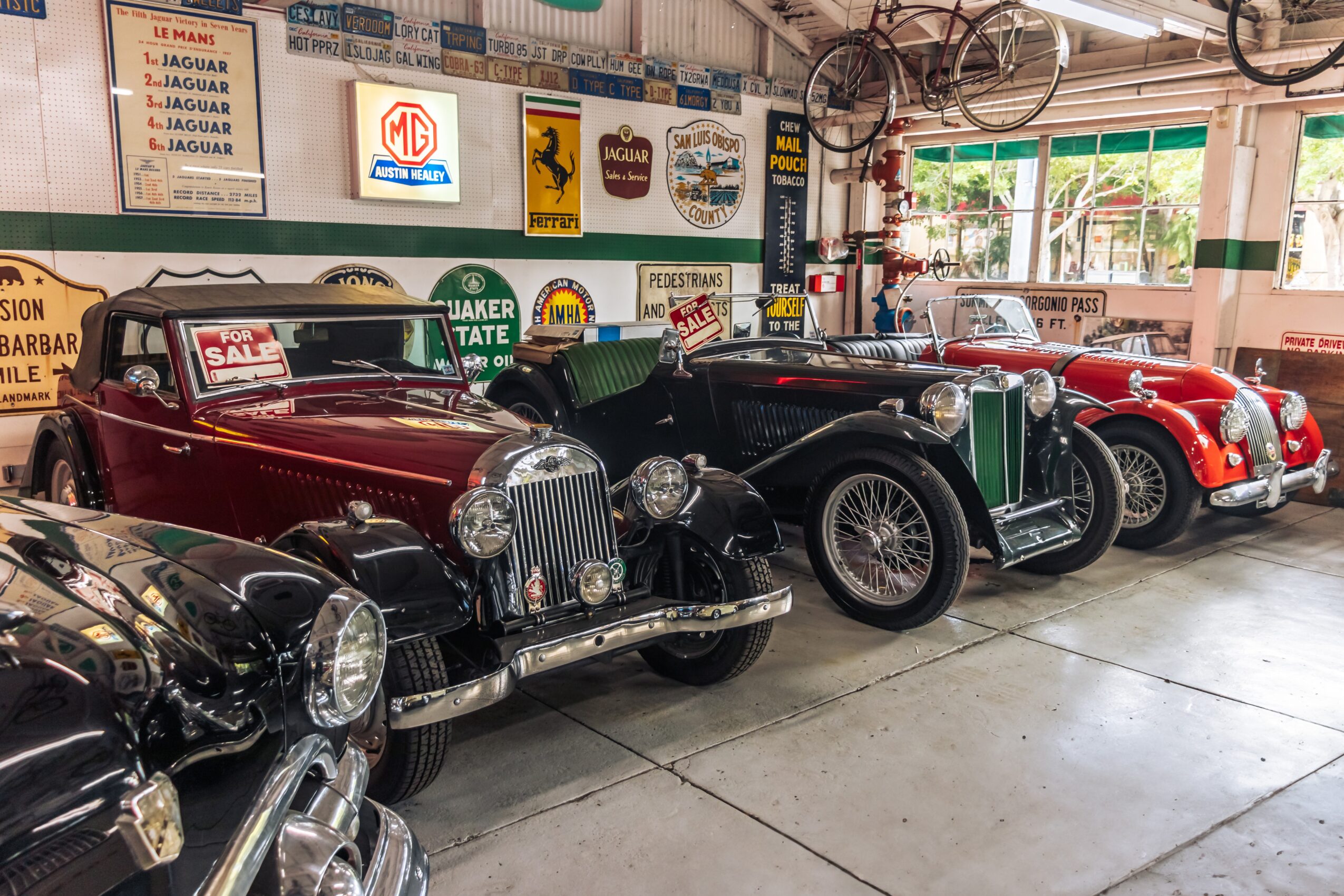 Four vintage cars parked indoors, with signs and banners on the wall behind them, and a window with green trim.