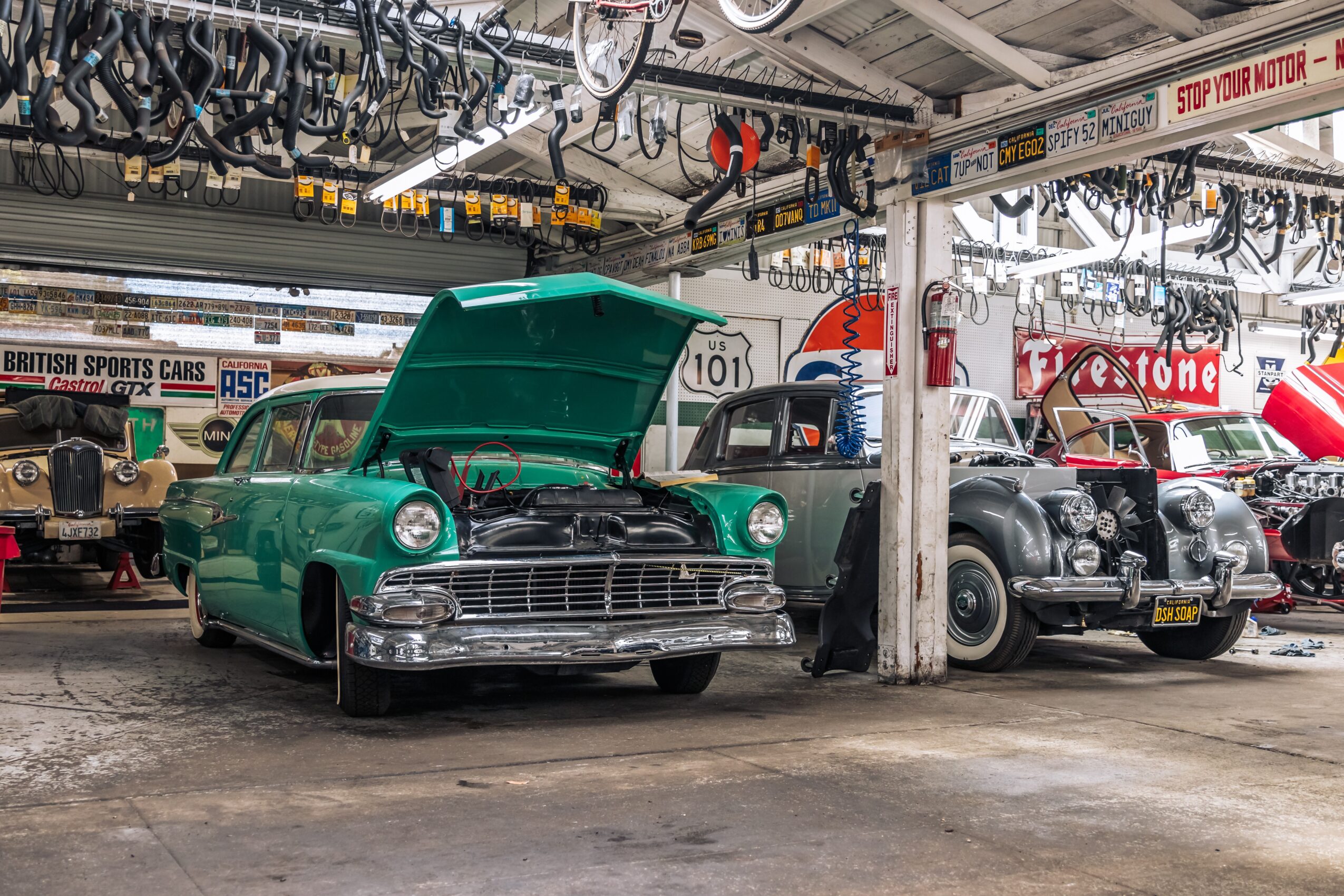 Green vintage car with open hood parked inside a garage alongside other classic cars, bicycles hanging from ceiling, and signs in background.