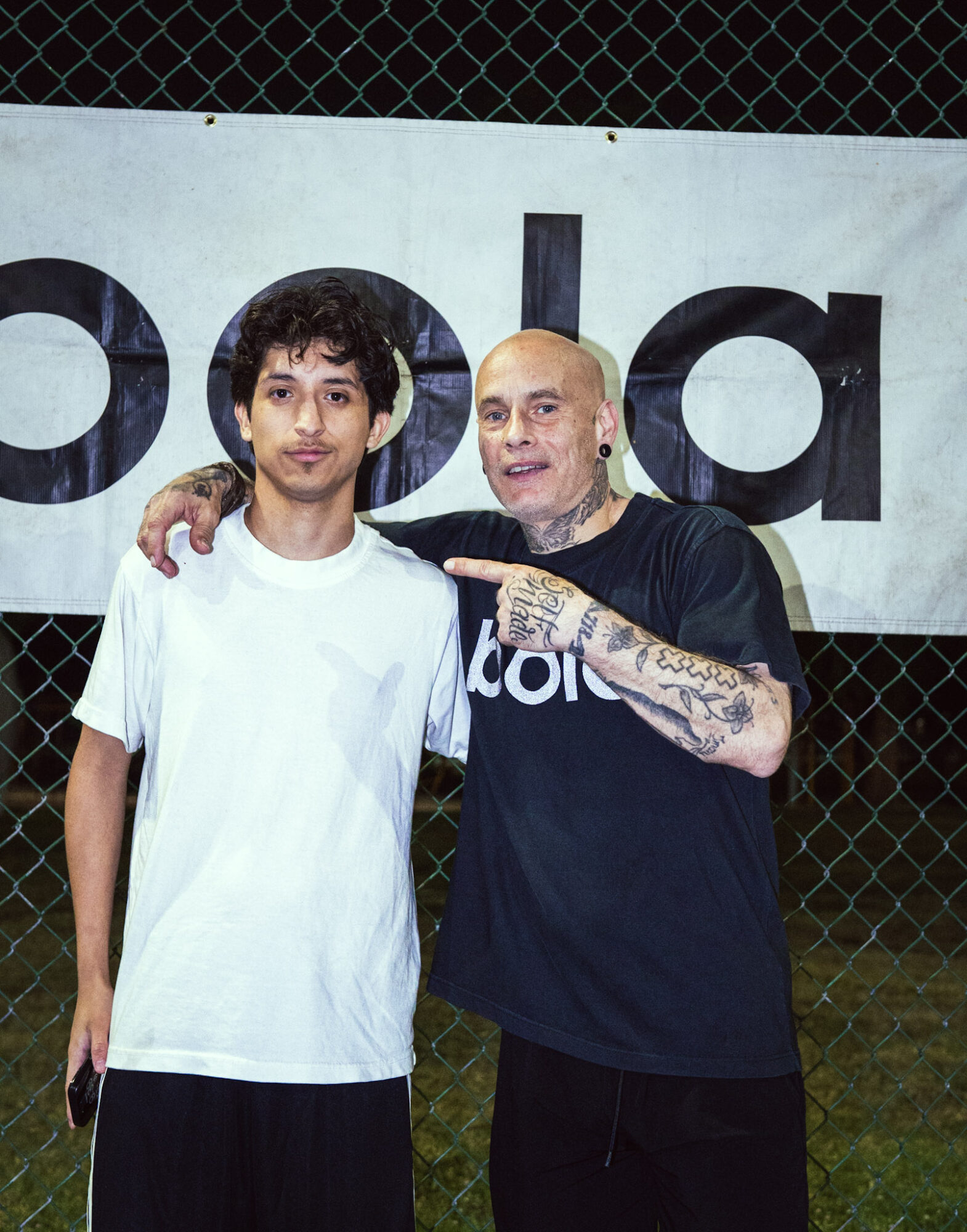 Two men standing outdoors in front of a banner, one with arm around the other, smiling at the camera.