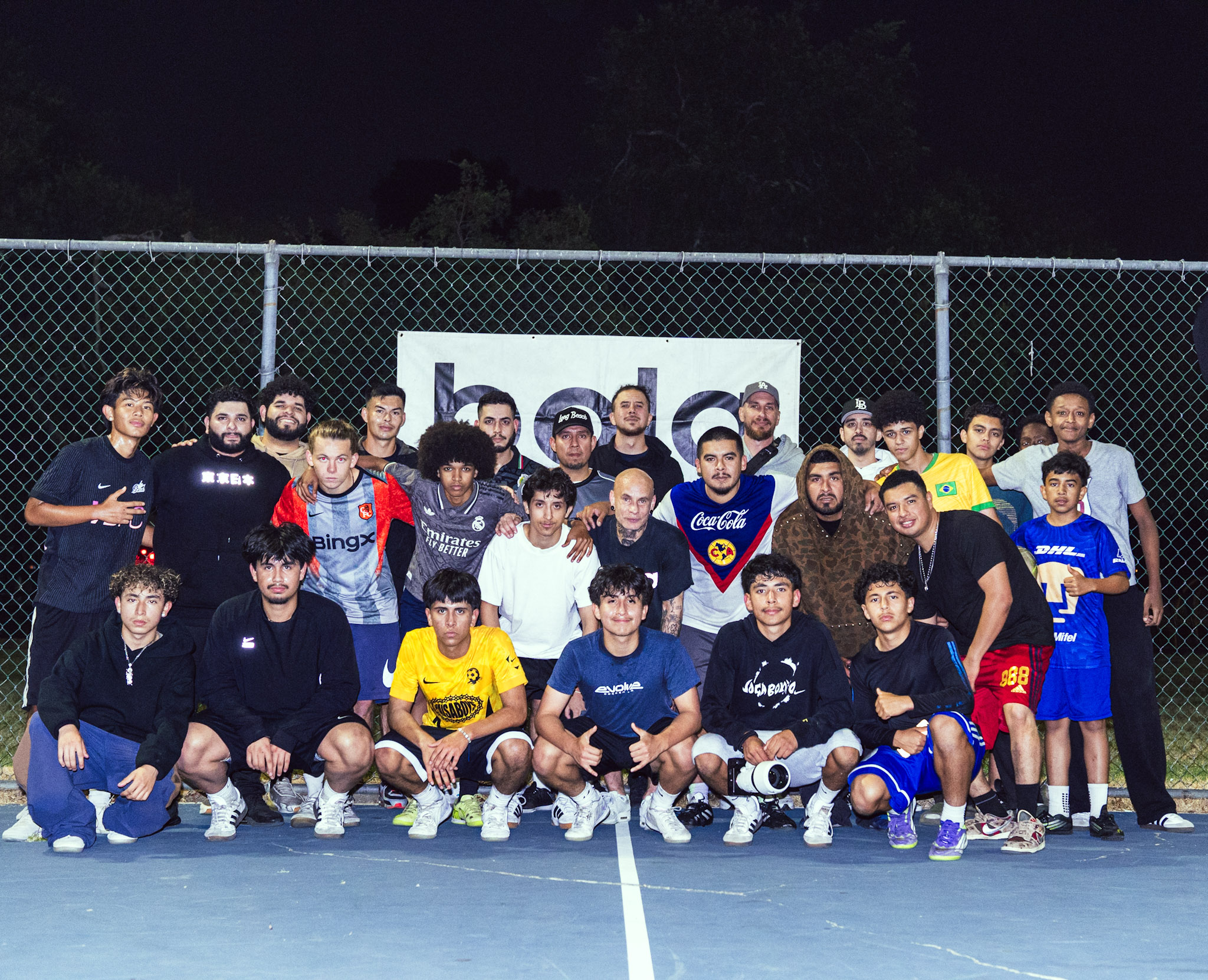 Group of young people and adults on a tennis court at night, posing for a photo in front of a fence and banner.
