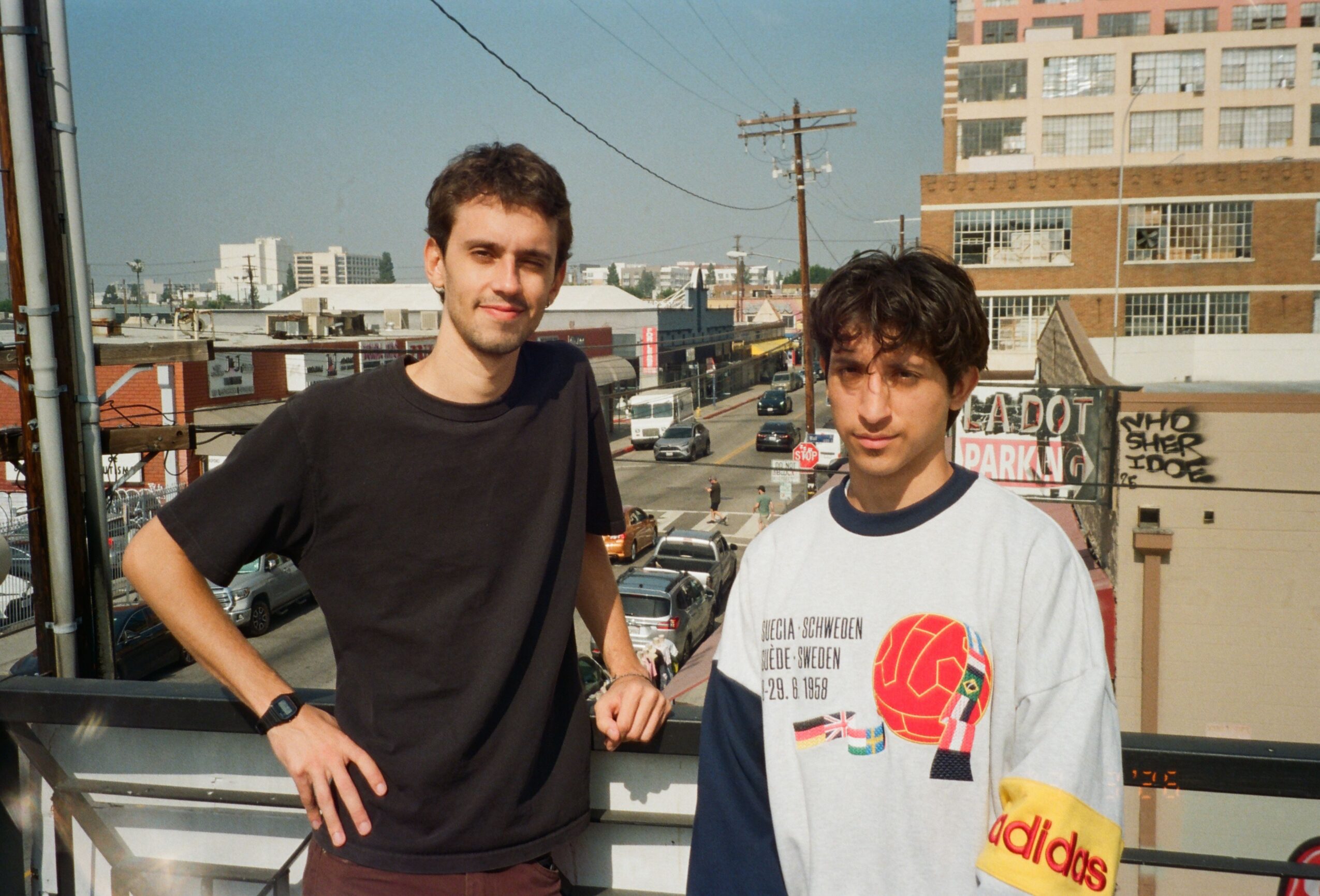 Two young men stand on a balcony with city buildings and power lines in the background, smiling at the camera.