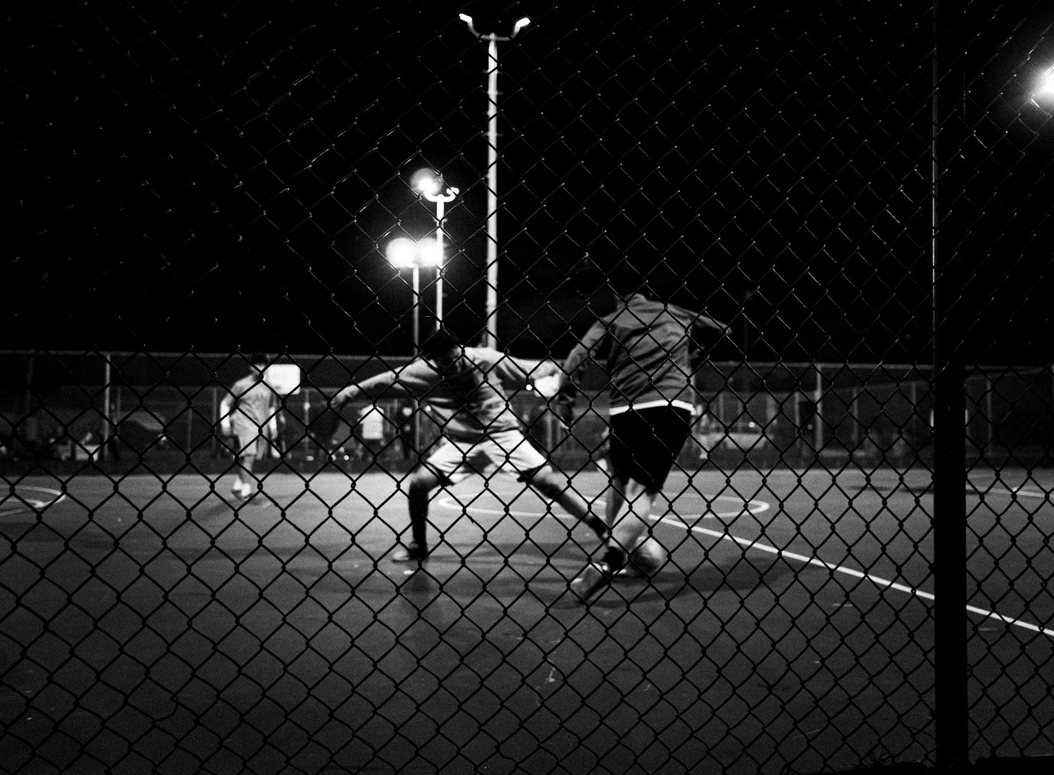 Two people playing tennis at night on a court with bright lights, viewed through a chain-link fence.