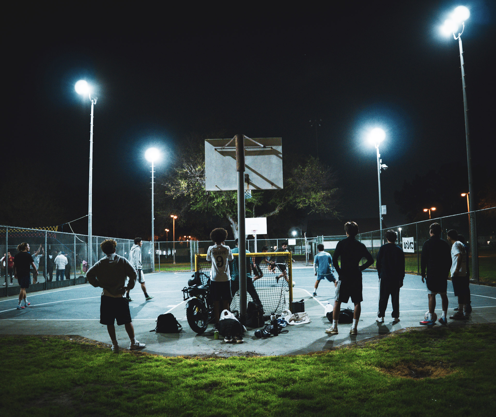 People gather around a basketball hoop on an outdoor court at night, illuminated by tall bright lights, with others in the background.