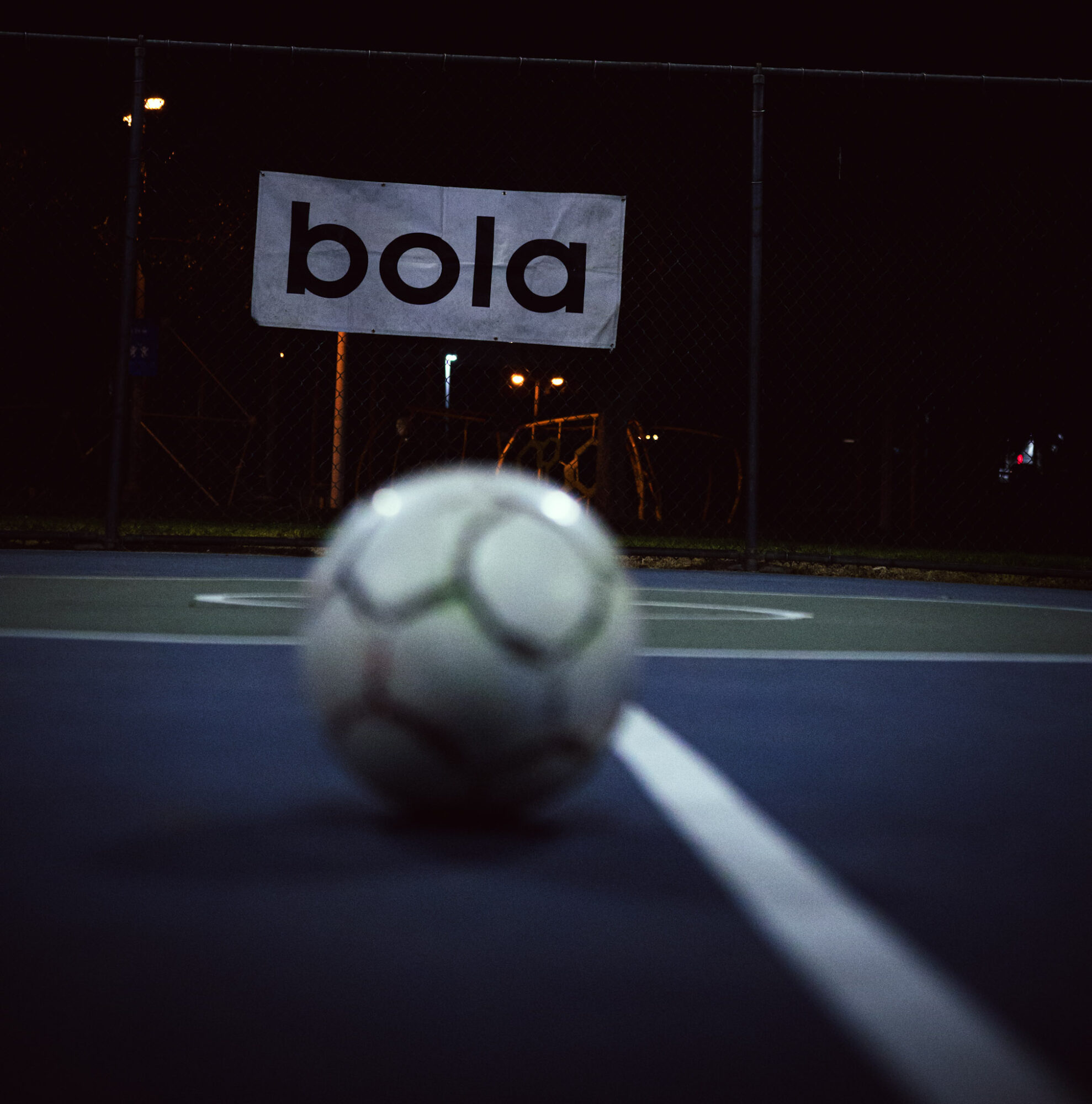 Soccer ball on a court at night with a sign reading 'bola' in the background.