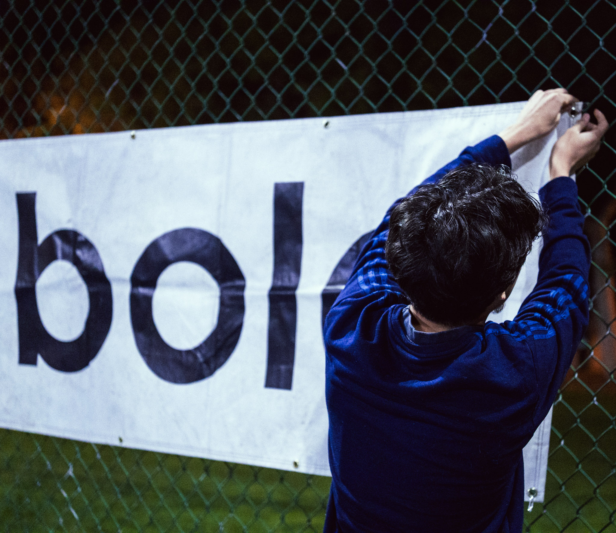 Person attaching a banner to a fence, with the banner displaying partially visible text, outdoors at night.