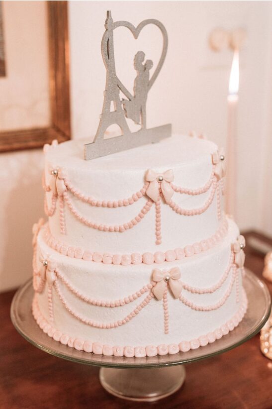 Two-tiered wedding cake with pink and white decorations, topped with a silver heart-shaped cake topper featuring a couple silhouette.