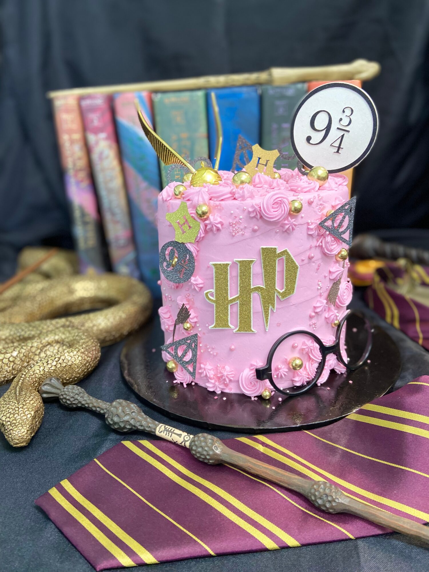 Pink Harry Potter-themed cake with gold initials, glasses, and a wand, surrounded by books and a striped cloth.