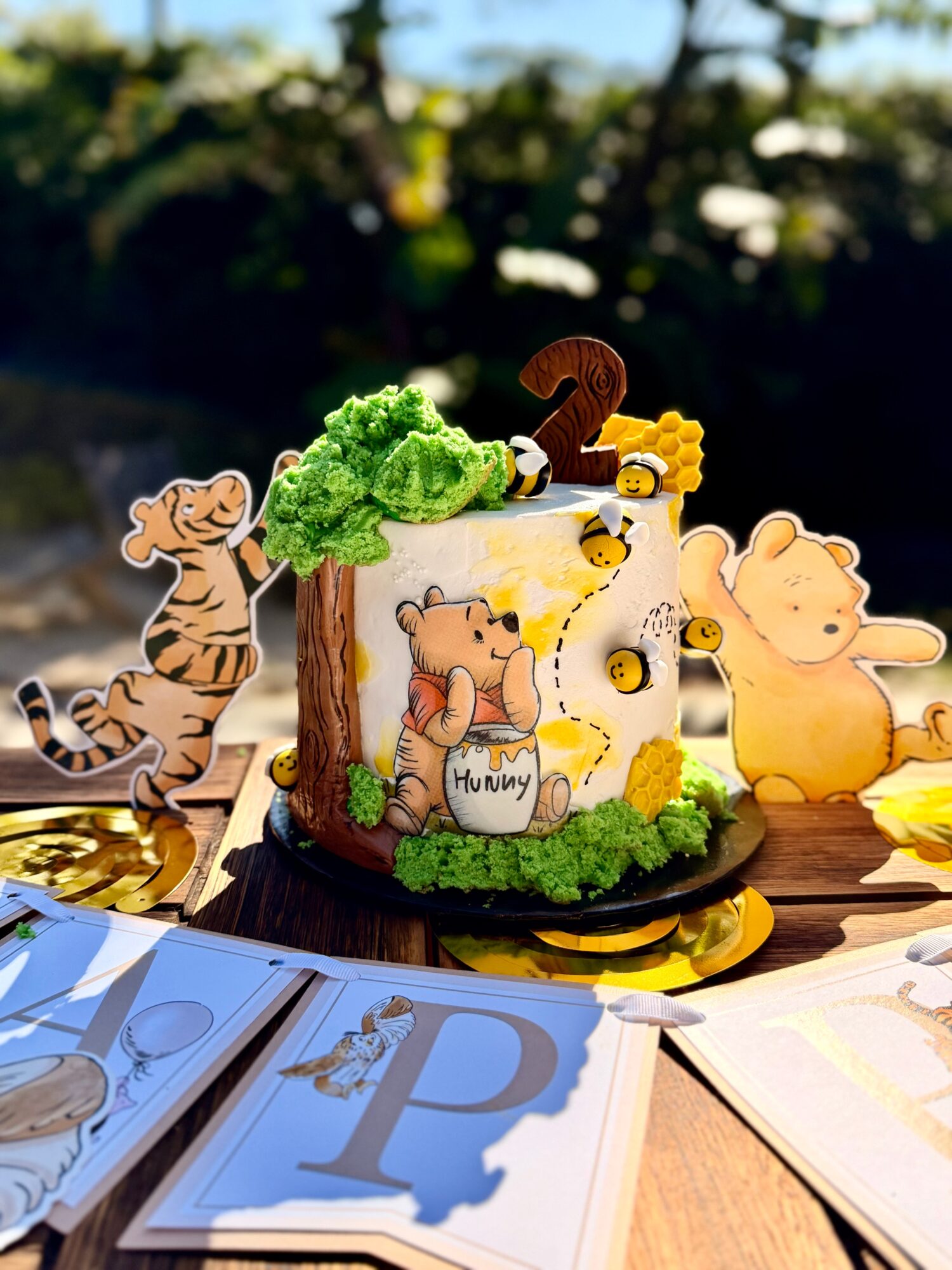 Decorative cake with animal figures and greenery, celebrating a child's second birthday outdoors, with papers and a table surface visible.